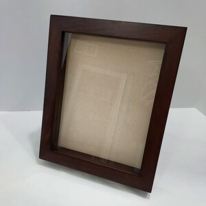 May include: A rectangular picture frame with a dark brown wood finish and a clear glass front. The frame features a deep profile design with a light beige, speckled mat board insert visible inside the central opening of the display area.