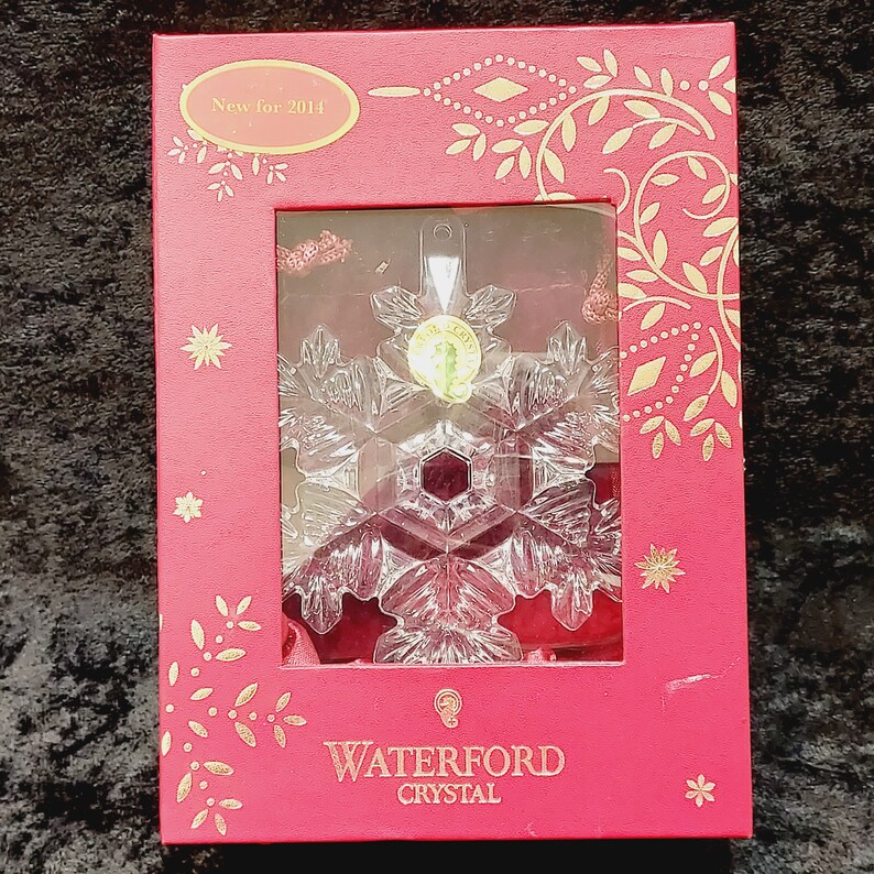 K&ouml;nnte beinhalten: Eine rote Schachtel mit goldenen Akzenten und einem klaren Kristall-Schneeflocken-Anh&auml;nger im Inneren. Die Schachtel tr&auml;gt den Text "New for 2014" und "Waterford Crystal".