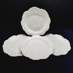 May include: A set of four white ceramic plates featuring a raised, embossed leaf pattern on the surface. Each plate has a scalloped, irregular edge that mimics the natural shape of a leaf, with a smooth, glossy finish across the entire set.