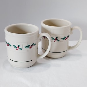 May include: Two off-white ceramic mugs featuring a repeating pattern of green holly leaves and red berries around the center. Each mug has a smooth handle, a slightly flared rim, and a single thin dark green horizontal line near the base.