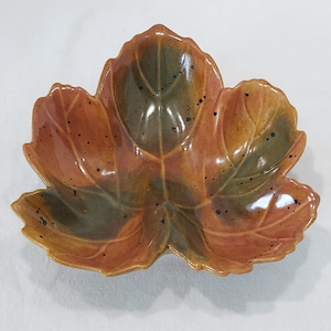 May include: A ceramic bowl shaped like a three-lobed leaf with a glossy finish. The surface features a blend of earthy orange and muted green tones, accented with dark brown speckles throughout the glaze and embossed vein details.