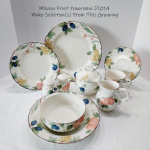 May include: A collection of Mikasa Fruit Panorama DC014 tableware. The set includes plates, bowls, cups, a creamer, a sugar bowl, and salt and pepper shakers. Each piece is white with a fruit design featuring plums, peaches, and grapes.