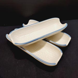 May include: Three white ceramic corn on the cob serving dishes with blue trim. The dishes are stacked on top of each other.