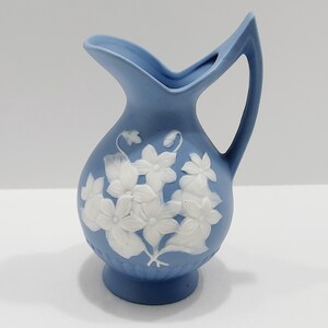 May include: A small, light blue ceramic pitcher with a handle and a spout. The pitcher is decorated with white floral relief designs. The pitcher is approximately 5 inches tall.
