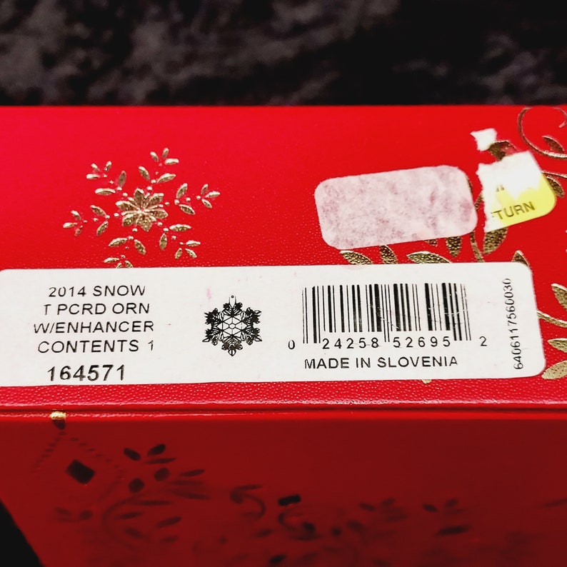 K&ouml;nnte beinhalten: Eine rote Box mit einem wei&szlig;en Etikett, auf dem "2014 SNOW T PCRD ORN W/ENHANCER CONTENTS 1 164571" steht. Das Etikett enth&auml;lt auch eine Schneeflocke und einen Barcode. Die Box ist in Slowenien hergestellt.