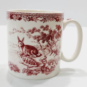 May include: A cream-colored ceramic mug featuring a dark red transferware pattern. The design depicts a rabbit and a turtle in a landscape with trees, bordered by a decorative floral band at the top rim. The mug has a curved handle on the right side.