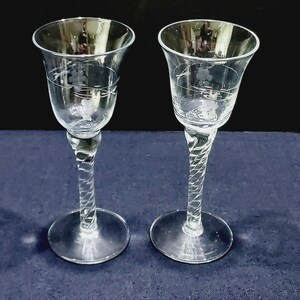 May include: Two clear, stemmed wine glasses. Each glass features a flared bowl with etched floral designs and a twisted stem. The base is round and flat. The glasses are displayed on a dark blue surface.