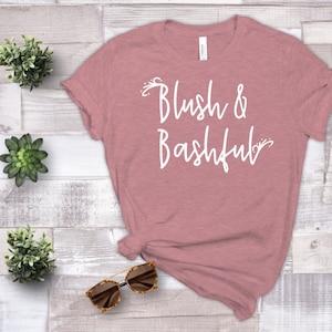 May include: A dusty rose coloured t-shirt with the text "Blush & Bashful" in white script.