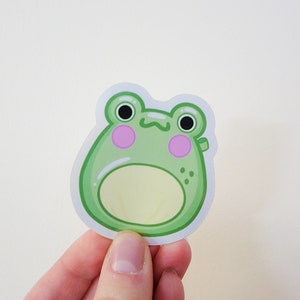 May include: A green frog sticker with a light yellow belly and pink cheeks. The frog has black eyes and a small smile.