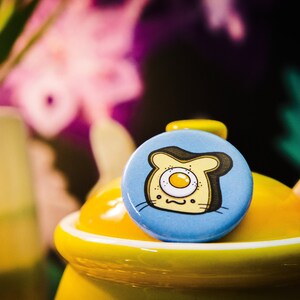 May include: A blue button with a cartoon illustration of a slice of bread with a fried egg inside. The bread has a cute face with a single eye and a smile. The button is sitting on top of a yellow ceramic jar with a wooden spoon.