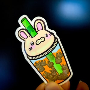 May include: A sticker of a cartoon bunny with a green straw sticking out of its head. The bunny is inside a clear cup filled with orange liquid and brown boba pearls.