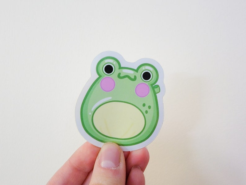 May include: A green frog sticker with a light yellow belly and pink cheeks. The frog has black eyes and a small smile.
