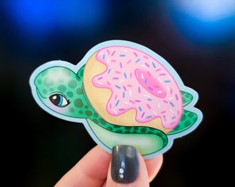 May include: A green sea turtle with a pink and white donut on its back. The donut has sprinkles on top.