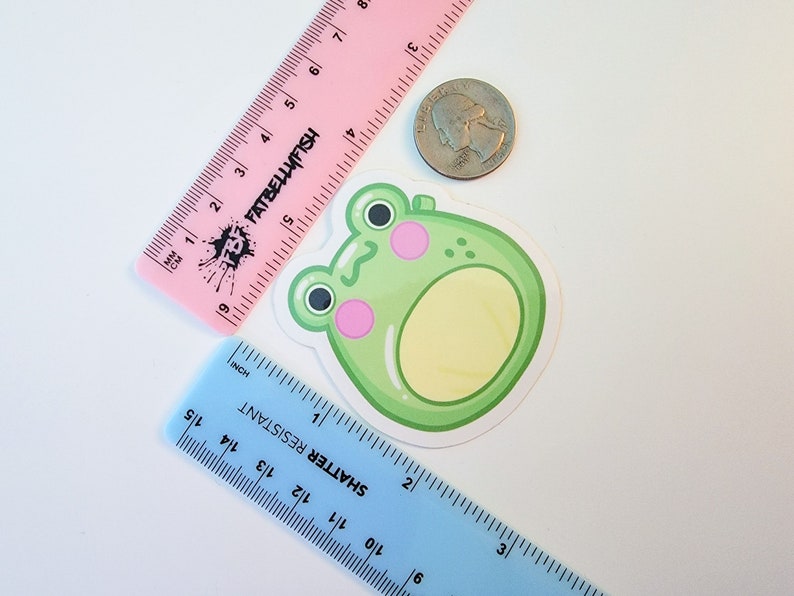 May include: A green frog sticker with pink cheeks and a yellow belly. The sticker is about 2 inches in diameter and is on a white background.