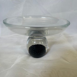 May include: A clear glass pedestal with a round, black glass sphere in the center. The pedestal is designed to hold a plate or other object.