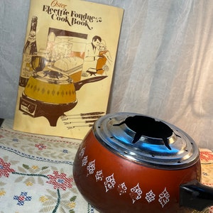 May include: Vintage Oster Electric Fondue Cook Book with a brown and white fondue pot with a black handle. The cookbook features a couple enjoying fondue with a bottle of wine.