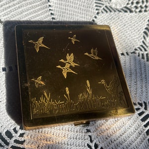 May include: A gold-toned metal compact with an engraved design of birds flying over a field of grass. The compact has a hinged lid and a small latch.