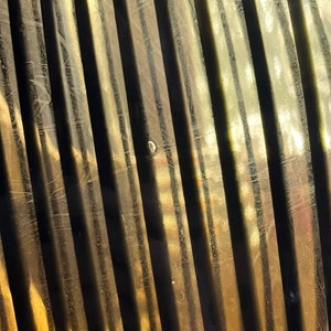 Op de afbeelding: Een close-up van een getextureerd, goudkleurig oppervlak met verticale lijnen. Het oppervlak is licht gebogen en heeft een subtiele glans.