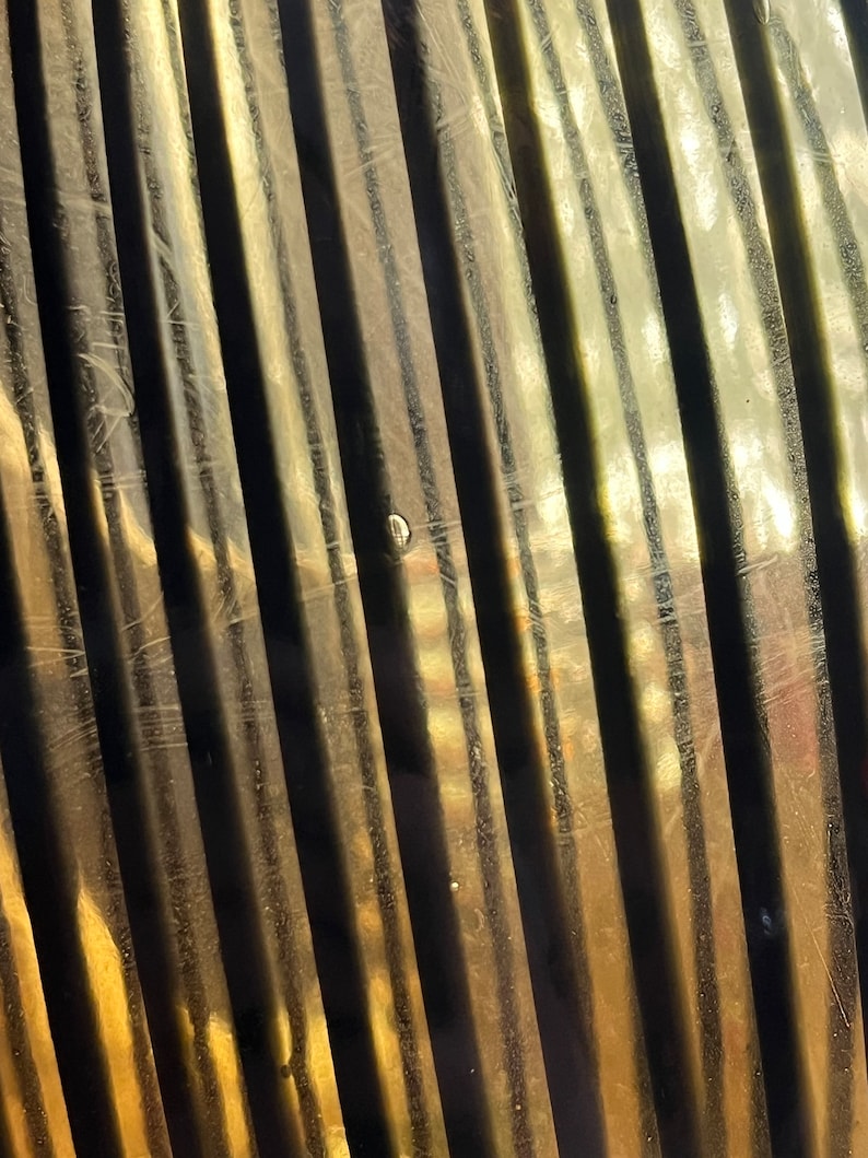 Op de afbeelding: Een close-up van een getextureerd, goudkleurig oppervlak met verticale lijnen. Het oppervlak is licht gebogen en heeft een subtiele glans.