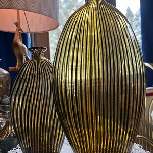 Op de afbeelding: Twee glazen vazen met zwarte en gouden streepjespatronen. De grotere vaas is groter en breder dan de kleinere vaas.