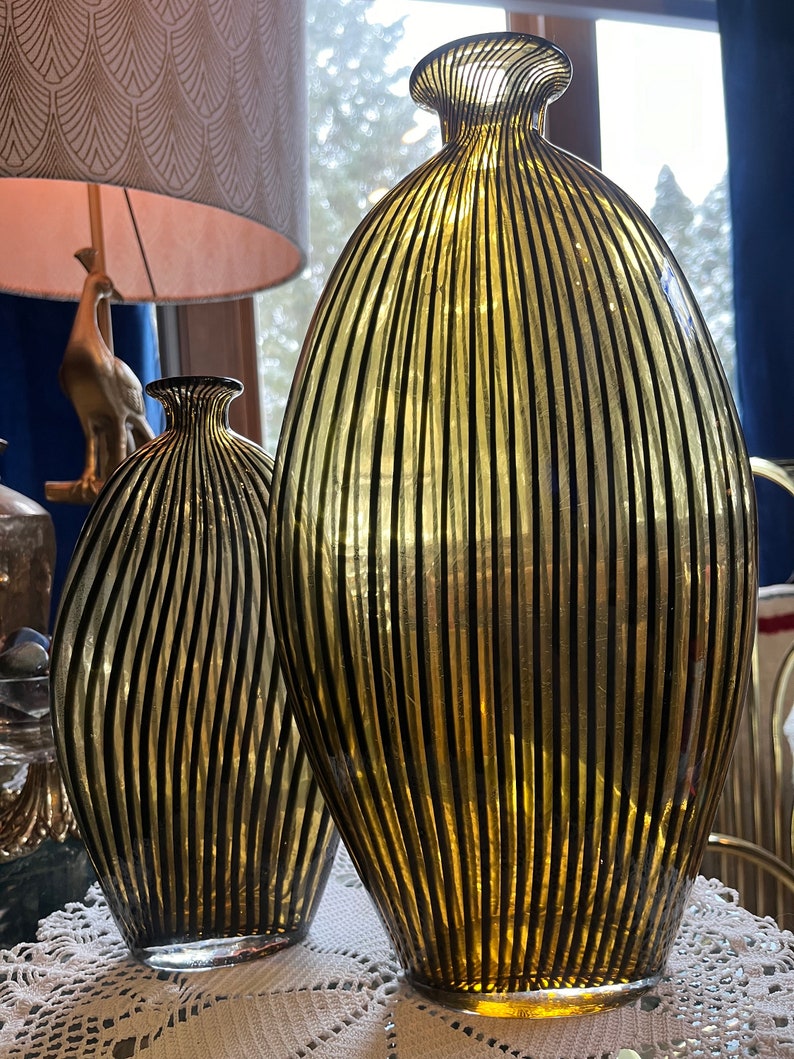 Op de afbeelding: Twee glazen vazen met zwarte en gouden streepjespatronen. De grotere vaas is groter en breder dan de kleinere vaas.