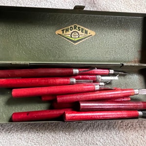 May include: An open, olive green metal toolbox containing several wood carving tools. The tools have red wooden handles and silver metal blades. The toolbox has a label that reads "THORSEN".