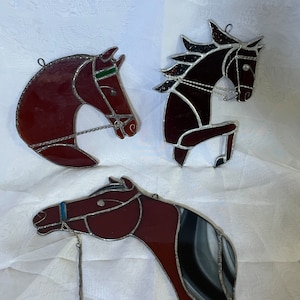May include: Three stained glass horse head ornaments. The horses are brown with black and white accents. The horses have silver reins and bridles.