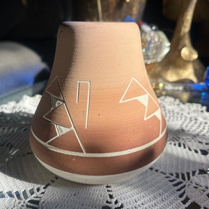 Può includere: Un piccolo vaso in ceramica marrone con un design geometrico bianco. Il vaso ha un fondo arrotondato e un'ampia apertura nella parte superiore.