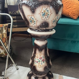 May include: A white and brown ceramic planter with floral designs sits on a matching pedestal stand. The planter has a scalloped rim and intricate details. The stand has a round base and a fluted column.