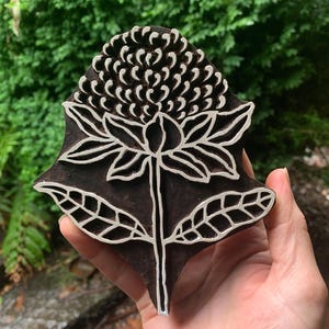 May include: A wooden block printing stamp featuring a detailed design of a flower with leaves. The flower has a large, round centre with petals radiating outwards. The leaves are long and narrow.