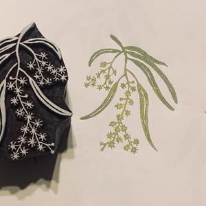 May include: A dark-coloured wooden printing block featuring a carved botanical design of a wattle branch with leaves and small flowers, accompanied by two green ink impressions of the same floral pattern stamped on a light-coloured surface.