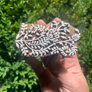 May include: A wooden block printing stamp featuring a white floral design. The stamp is carved with intricate details of leaves and flowers.