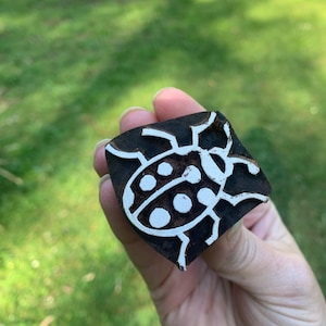 May include: A square wooden block featuring a white ladybird design. The ladybird has a rounded body with white spots and a detailed outline. The block is held against a blurred green background.