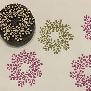 May include: A wooden block printing stamp with a white design of a wreath of leaves and berries. There are three prints made with the stamp, one in green and two in pink.