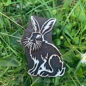 May include: A dark brown, rectangular stamp featuring a white rabbit design. The rabbit is depicted in a seated position. The stamp is set on green grass, with small white daisies scattered around.