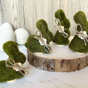 May include: Four green moss bunny figurines with a brown twine bow and a white button on a wooden slice.