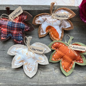 May include: Four handmade leaf ornaments with fabric patterns and stitched details. Each leaf has a small tag with words like "Gather," "Thankful," "Harvest," and "Autumn." The leaves are in shades of red, orange, brown, and beige, with a rustic, autumnal theme.
