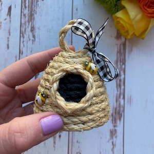 Puede incluir: Un pequeño adorno con forma de colmena hecho de cuerda natural con una entrada pintada de negro y adornos de abejas amarillas y negras. El adorno está atado con una cinta a cuadros en blanco y negro.