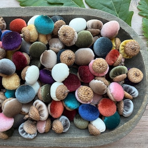 May include: A collection of decorative acorns featuring soft velvet fabric caps in a wide variety of colors including red, blue, green, yellow, pink, and neutral tones. Each acorn has a textured, natural-looking brown cap attached to the top.