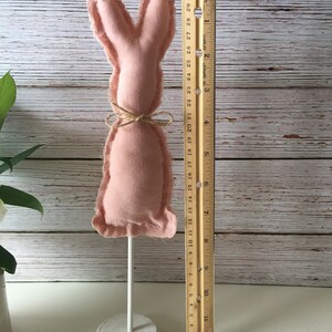 May include: A pink fabric bunny rabbit with a brown twine bow, standing on a white wooden base. The bunny is approximately 6 inches tall.