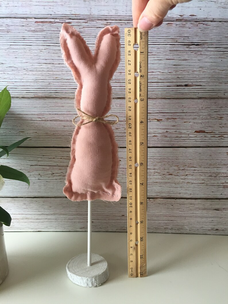 May include: A pink fabric bunny rabbit with a brown twine bow, standing on a white wooden base. The bunny is approximately 6 inches tall.