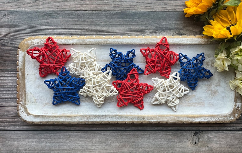 May include: A tray of woven star decorations in red, white, and blue. The stars are arranged in a row on a white tray with a gold trim.
