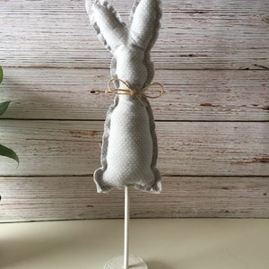 May include: A gray fabric bunny with white polka dots, tied with twine, stands on a white wooden base.