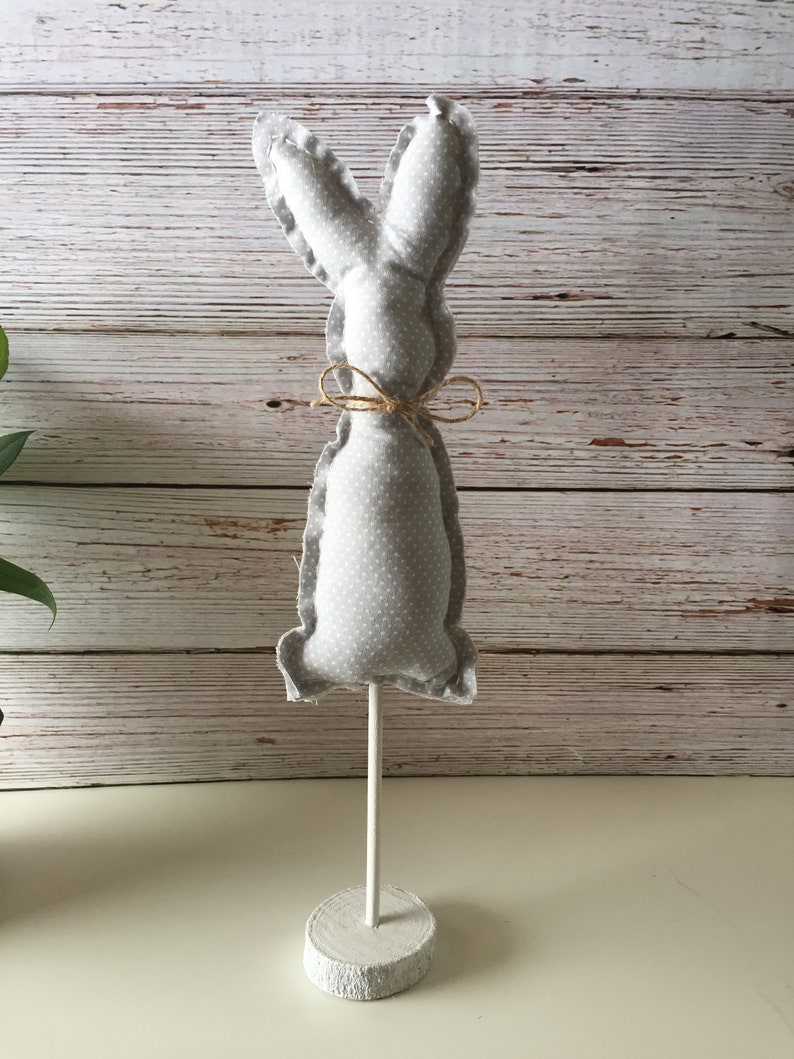May include: A gray fabric bunny with white polka dots, tied with twine, stands on a white wooden base.