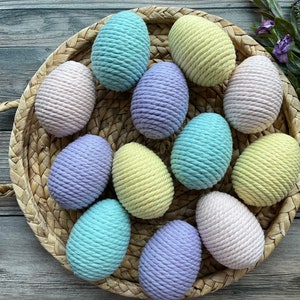 Op de afbeelding: Een rieten mand gevuld met pastelkleurige touw-paaseieren. De eieren zijn in tinten geel, roze, paars en blauw.
