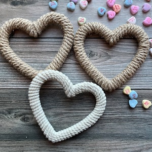 May include: Three rope hearts, two brown and one white, are arranged on a wooden surface.  The hearts are made of thick rope and are tied together in the center.  The background is a rustic wooden surface with scattered candy hearts.