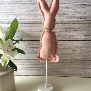 May include: A pink fabric bunny rabbit with a brown twine bow around its neck. The bunny is attached to a white wooden stand.