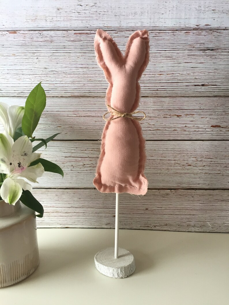 May include: A pink fabric bunny rabbit with a brown twine bow around its neck. The bunny is attached to a white wooden stand.
