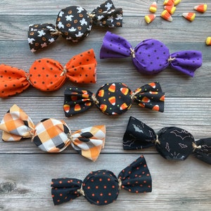 May include: Eight Halloween-themed fabric candy wrappers with different patterns, including ghosts, bats, polka dots, and candy corn. The wrappers are tied with twine and are arranged on a wooden surface.