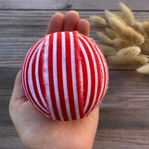 Puede incluir: Una bola de adorno de tela a rayas rojas y blancas. La bola est&aacute; hecha a mano y tiene una superficie ligeramente irregular.
